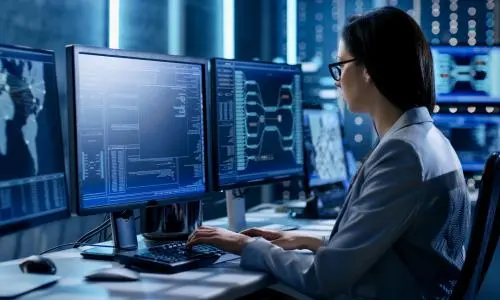 A woman with black hair and fair skin sitting at a desk with three computer screens in front of her